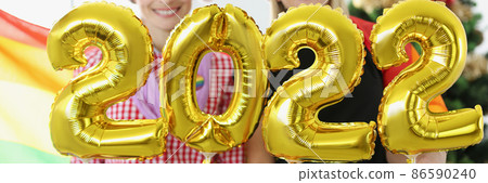 Two women in santa hat holding helium balloons with numbers 2022 on background of lgbt flag and Christmas tree closeup Two women in santa hat holding helium balloons with numbers 2022 on background of lgbt flag and Christmas tree closeup 86590240