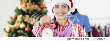 Seamstress in red Santa hat holding alarm clock and scissors near Christmas tree Seamstress in red Santa hat holding alarm clock and scissors near Christmas tree 86590242
