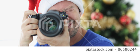 Man in red santa hat taking pictures with camera near christmas tree Man in red santa hat taking pictures with camera near christmas tree 86590244
