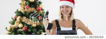 Woman builder in red santa claus hat holding drill in hands near christmas tree Woman builder in red santa claus hat holding drill in hands near christmas tree 86590269