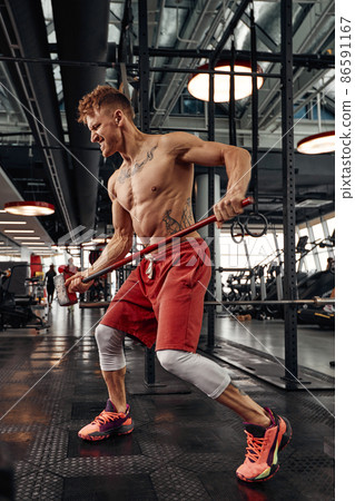 Strength concept. Male athlete using hammer during fitness workout. Fitness man beating rubber tire in gym. Muscular sportsman performing intensity training with sledgehammer in sport club 86591167