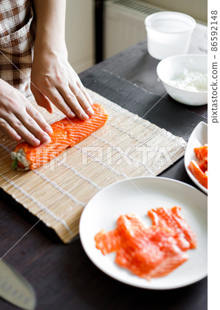 Young woman wrapping up sushi roll ingredients on bamboo mat. Young woman wrapping up sushi roll ingredients on bamboo mat. 86592148