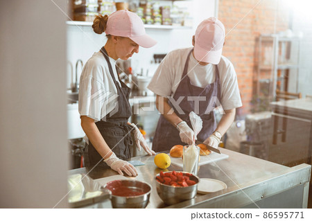 Young womancuts bun with colleague at metal counter in craft bakery shop Young womancuts bun with colleague at metal counter in craft bakery shop 86595771