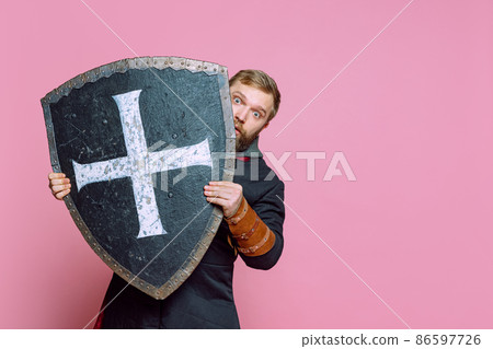 Portrait of scary man, medieval warrior or archer in armor hiding himself behind a shield isolated over pink studio background Portrait of scary man, medieval warrior or archer in armor hiding himself behind a shield isolated over pink studio background 86597726