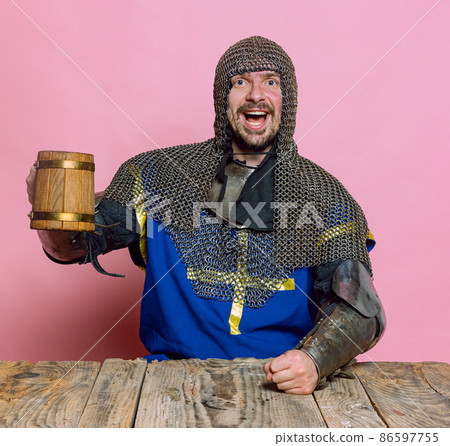 Portrait of excited man, medieval warrior, knight in special protection rising wooden beer mug isolated over pink studio background 86597755