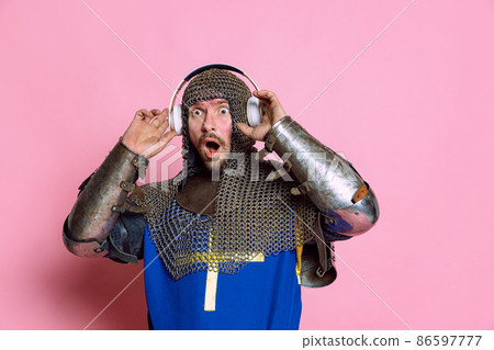 Half-length portrait of man, medieval warrior or knight listening to music in headphones with shocked expression isolated over pink background 86597777