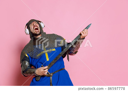 Half-length portrait of man, medieval warrior or knight in armor holding sword like guitar isolated over pink background Half-length portrait of man, medieval warrior or knight in armor holding sword like guitar isolated over pink background 86597778