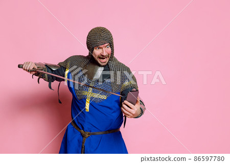 Half-length portrait of man, medieval warrior or knight in armor pointing sword at phone screen isolated over pink studio background 86597780