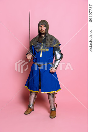 Full-length portrait of serious looking man, brtutal medieval warrior or knight in protective covering with sword isolated over pink studio background Full-length portrait of serious looking man, brtutal medieval warrior or knight in protective covering with sword isolated over pink studio background 86597787