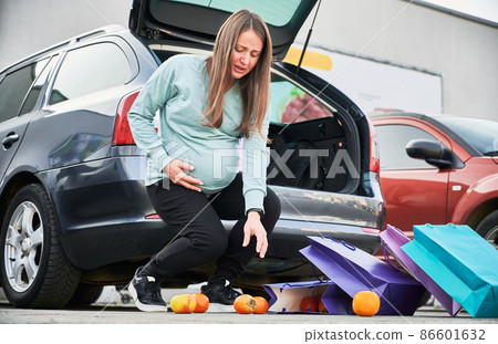Woman in third trimester of pregnancy trying to pick up oranges from the ground, which fell from shopping bags, supporting her belly with her hand. 86601632