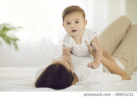 Portrait of happy baby boy cuddling together with his mommy on bed at home. Young mother and adorable little child in comfy white cotton home wear lying on bed in the morning. Family and care concept 86603232