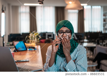 Smiling young woman talk on smartphone work on laptop look in distance thinking. Happy African American customer client use modern gadgets cellphone and computer at home office. Smiling young woman talk on smartphone work on laptop look in distance thinking. Happy African American customer client use modern gadgets cellphone and computer at home office. 86603531