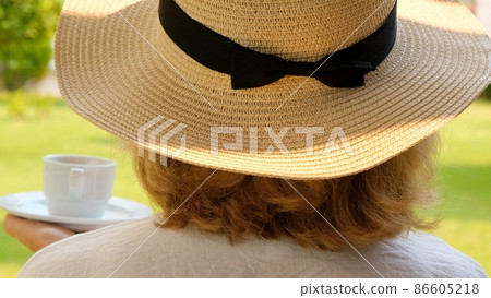 Back view of a woman in a straw hat with a white cup of hot drink of coffee in her own garden on the 86605218