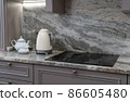 beautiful closeup of custom designed kitchen, with marble looking quartz countertop and backsplash. cream electric kettle with porcelain tea accessories on the marble countertop next to ceramic hob 86605480