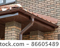 Corner of house with new brown metal tile roof and rain gutter. Metallic Guttering System, Guttering and Drainage Pipe Exterior 86605489