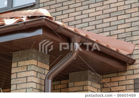 Corner of house with new brown metal tile roof and rain gutter. Metallic Guttering System, Guttering and Drainage Pipe Exterior Corner of house with new brown metal tile roof and rain gutter. Metallic Guttering System, Guttering and Drainage Pipe Exterior 86605489