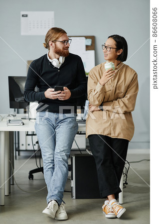 Two young intercultural businessmen in casual clothes standing by desk Two young intercultural businessmen in casual clothes standing by desk 86608066