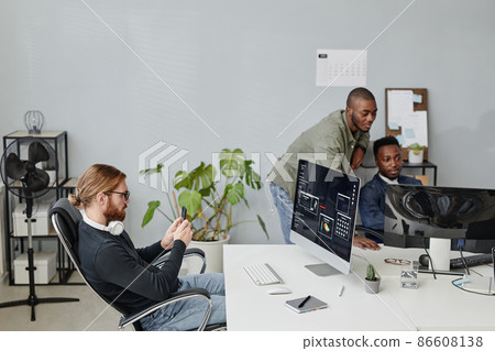 Young businessman with smartphone sitting by desk in front of computer screen Young businessman with smartphone sitting by desk in front of computer screen 86608138