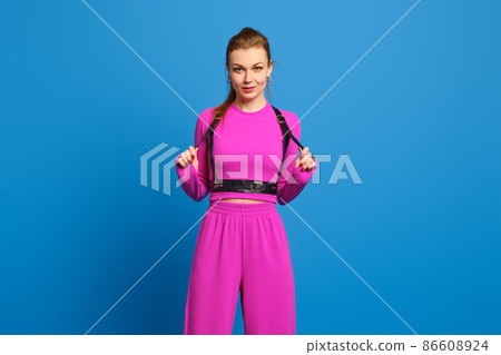 Trendy woman in purple knitted pantsuit holds the belts of leather shoulder harness over blue studio background Trendy woman in purple knitted pantsuit holds the belts of leather shoulder harness over blue studio background 86608924
