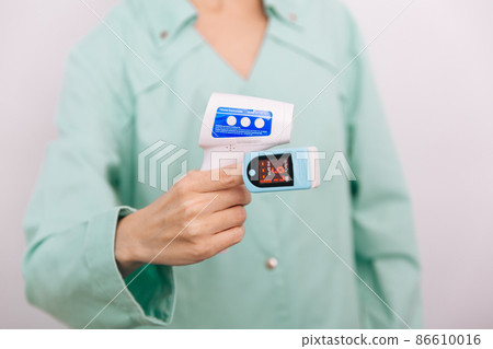 Temperature measurement gun in doctor hands. Close-up shot of doctor wearing protective surgical mask ready to use infrared isometric thermometer gun to check body temperature for virus symptoms. 86610016