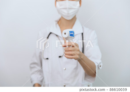 Temperature measurement gun in doctor hands. Close-up shot of doctor wearing protective surgical mask ready to use infrared isometric thermometer gun to check body temperature for virus symptoms. Temperature measurement gun in doctor hands. Close-up shot of doctor wearing protective surgical mask ready to use infrared isometric thermometer gun to check body temperature for virus symptoms. 86610039