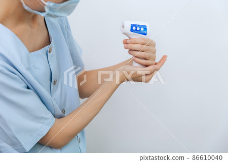 Temperature measurement gun in doctor hands. Close-up shot of doctor wearing protective surgical mask ready to use infrared isometric thermometer gun to check body temperature for virus symptoms. Temperature measurement gun in doctor hands. Close-up shot of doctor wearing protective surgical mask ready to use infrared isometric thermometer gun to check body temperature for virus symptoms. 86610040