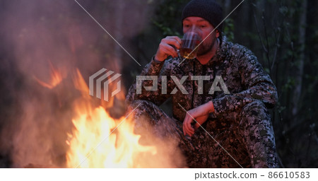 Man with cup of tea the fire in the forest. Human looks at the bonfire and drinks tea on the nature in the summer. Alone traveler sits and rests by the fire with a cup of coffee at dusk. Man with cup of tea the fire in the forest. Human looks at the bonfire and drinks tea on the nature in the summer. Alone traveler sits and rests by the fire with a cup of coffee at dusk. 86610583