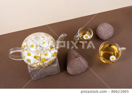 Chamomile tea. Cups of herbal tea, transparent teapot with camomile flowers and stones on brown background. Calming drink concept. Trendy still life 86621665