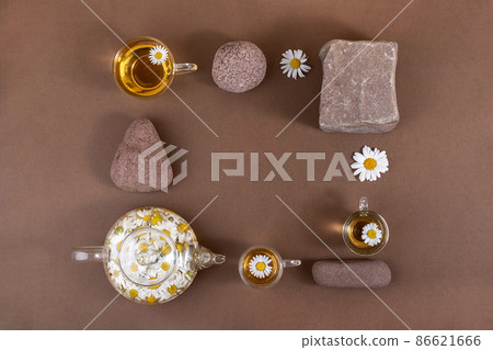 Chamomile tea. Cups of herbal tea, transparent teapot with camomile flowers and stones on brown background. Calming drink concept. Trendy still life Chamomile tea. Cups of herbal tea, transparent teapot with camomile flowers and stones on brown background. Calming drink concept. Trendy still life 86621666