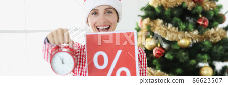 Young smiling woman in santa claus hat holds alarm clock and poster with percent on background of New Year tree Young smiling woman in santa claus hat holds alarm clock and poster with percent on background of New Year tree 86623507