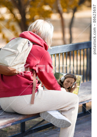 Video call. Woman in red hoodie sits on a bench and chatting with a friend using a laptop in the park outdoor in autumn, vertical photo, selected focus. Video call. Woman in red hoodie sits on a bench and chatting with a friend using a laptop in the park outdoor in autumn, vertical photo, selected focus. 86623697