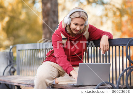 Student life. Caucasian woman sits on a bench using a laptop in the public park outdoor in autumn, selected focus. Student life. Caucasian woman sits on a bench using a laptop in the public park outdoor in autumn, selected focus. 86623698