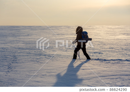 Lone human with backpack and ski poles walks througth snow-covered plain at sunset, selected focus. 86624290