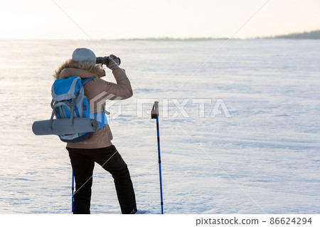 Human in beige down jacket with a blue backpack and ski poles looks through binoculars in the middle of a snowy plain in winter, back view, selected focus. 86624294