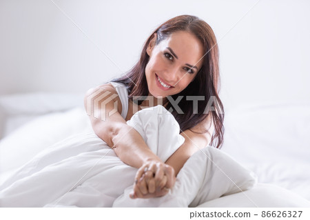 Good-looking woman sits covered in white feather-bed strecthing her arms, smiling Good-looking woman sits covered in white feather-bed strecthing her arms, smiling 86626327
