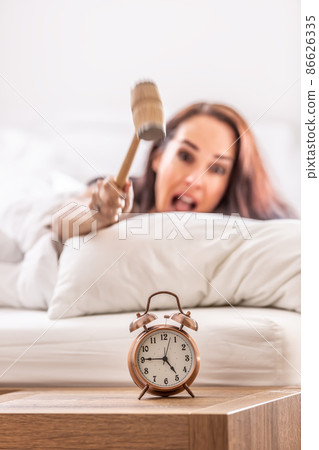 Smashing of alarm clock with the hammer by a woman reaching out from the bed Smashing of alarm clock with the hammer by a woman reaching out from the bed 86626335