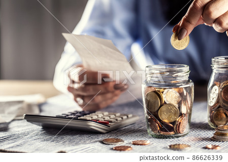 Detail of woman holding a bill in one hand and putting fifty cents into a glass jar with calculator on the table Detail of woman holding a bill in one hand and putting fifty cents into a glass jar with calculator on the table 86626339