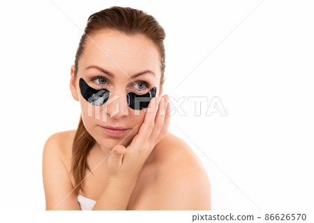 a woman applies cosmetic black patches under her eyes against a white wall 86626570