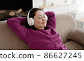 Young asian woman relaxing on comfortable sofa with eyes closed wearing headphones. Pretty mixed race lady enjoys listening chill music audio sound meditating feeling no stress at home. 86627245