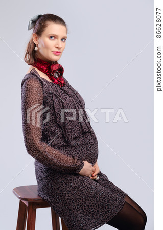 Pregnant woman in a loose dress sitting in profile on chair on grey studio background Pregnant woman in a loose dress sitting in profile on chair on grey studio background 86628077
