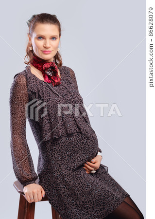Pregnant woman in a loose dress sitting in profile on chair on grey studio background Pregnant woman in a loose dress sitting in profile on chair on grey studio background 86628079