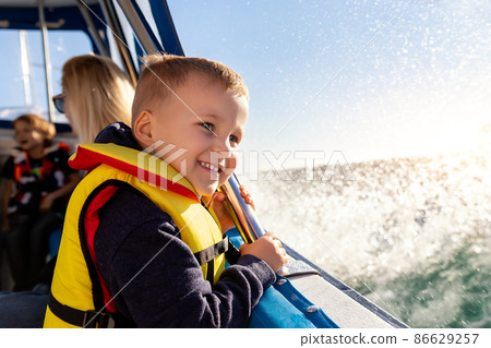 Portrait of cite little blond happy excited smiling caucasian boy wear lifevest enjoy sailing on motor boat sea against blue sky and water splash wave sun backlit. Summer travel vacation recreation 86629257