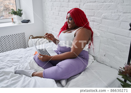 Multiracial red haired woman working at the laptop while sitting on the bed and chatting with somebody Multiracial red haired woman working at the laptop while sitting on the bed and chatting with somebody 86630271