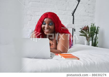 Red haired woman laughing while laying on the bed and working at the laptop Red haired woman laughing while laying on the bed and working at the laptop 86630274
