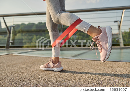 Close-up of the legs of an athlete doing body weight training with a resistance fitness elastic band on a city bridge. 86631007