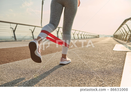 Rear view of the legs of an athlete woman, sportswoman in gray leggins doing lunges during body weight training, exercising with a resistance fitness elastic band on a city bridge at sunrise. Close-up 86631008