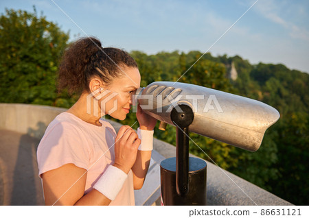 Woman looks through a telescope at the city. Athletic woman in a pink t-shirt is watching the beautiful green nature and the river background through stationary city binoculars 86631121