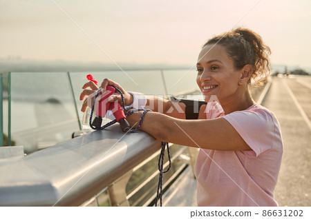 Confident relaxed middle aged sportswoman with a bottle of water and a skipping rope, admiring beautiful nature standing on the city bridge after intense cardio workout at sunrise 86631202