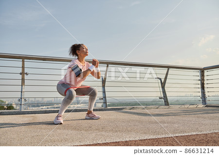 Determined middle aged sporty woman, female athlete exercising outdoor at sunrise, doing squats with elastic resistance band. Fitness sport endurance, body weight training and active lifestyle concept 86631214
