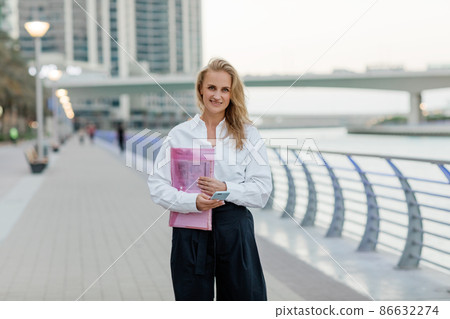 A beautiful business woman with a folder of documents in her hands outdoors. City portrait, business portrait A beautiful business woman with a folder of documents in her hands outdoors. City portrait, business portrait 86632274
